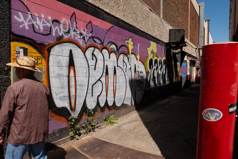 A person in a hat and brown shirt stands facing a graffiti-covered wall in an alley. The wall features large, colorful graffiti designs with the words "OBNOX." The alley has concrete ground and buildings on both sides, with shadows cast along the ground.