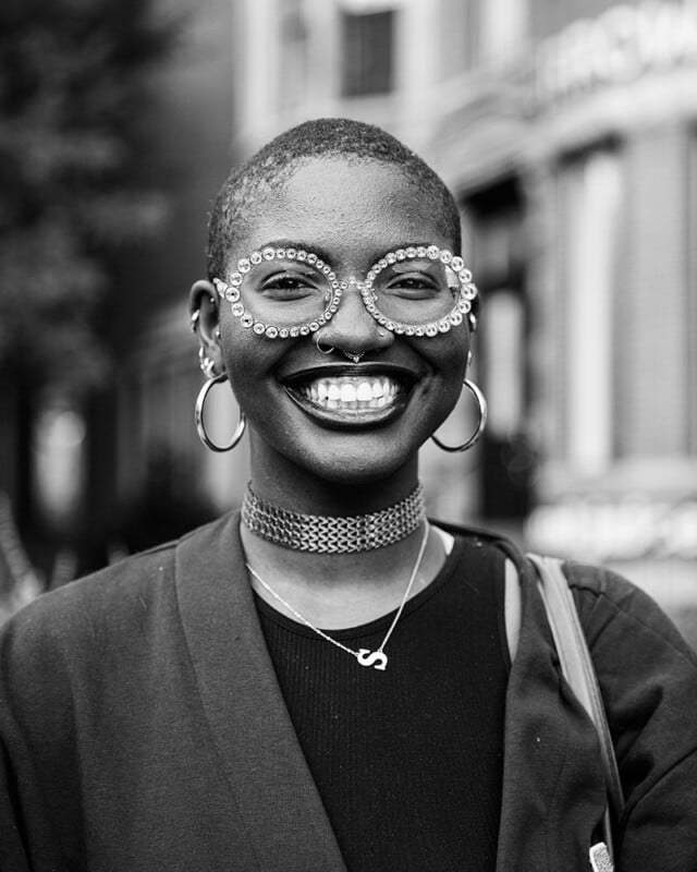 A person with closely cropped hair is smiling broadly. They are wearing large, round, rhinestone-studded glasses, hoop earrings, and a choker necklace. The background is blurred, featuring buildings and trees. The photo is in black and white.