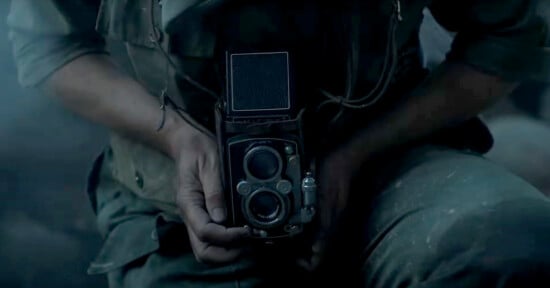 A person in worn clothing holds a vintage twin-lens reflex camera with care. The scene is dimly lit, evoking a feeling of nostalgia. The person's hands are prominently featured, revealing intricate details of the camera.