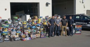 A group of five people, including police officers and civilians, stand in front of a large collection of boxed toys piled up outside a building labeled "Springfield Police". A police vehicle is parked nearby against the building's white wall.