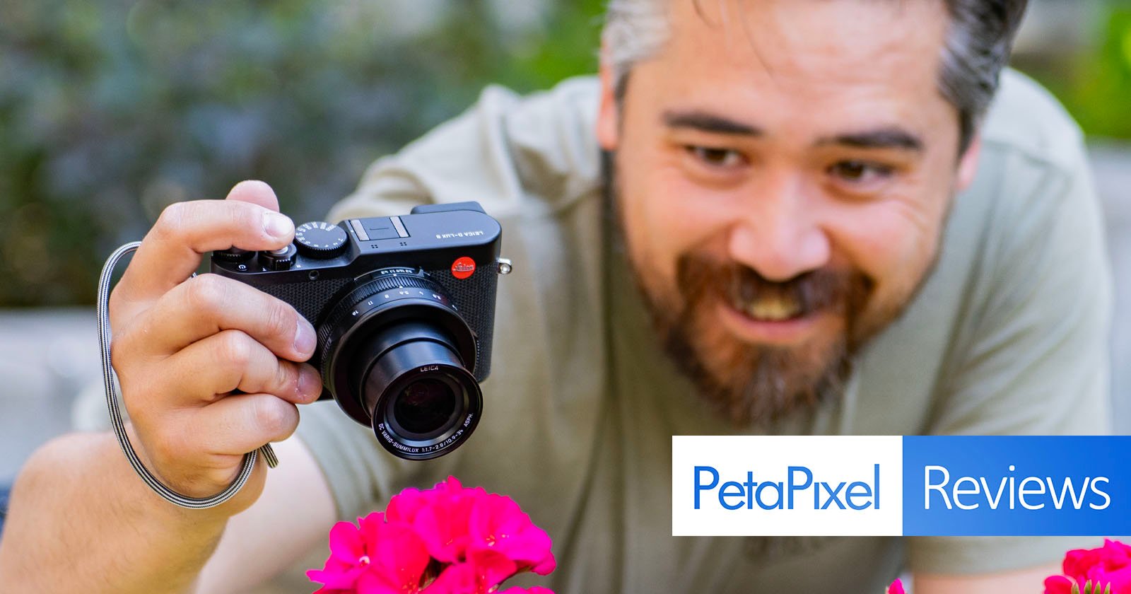 A man with a beard is smiling as he takes a close-up photo of pink flowers with a black camera. The background is blurred, highlighting the flowers and the photographer. There is an overlay with the text "PetaPixel Reviews" at the bottom right corner.