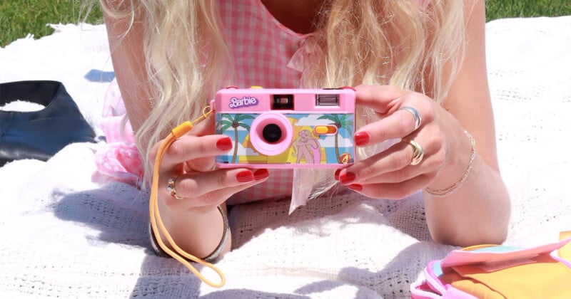 A person with long blonde hair, wearing pink checkered clothing, is holding a pink Barbie-themed camera. The person's fingernails are painted red and they are sitting on a white blanket outdoors. The background includes green grass and a glimpse of a pink bag.