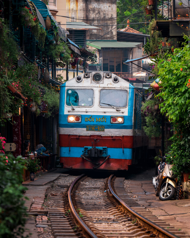 A blue and orange train travels down narrow tracks through the middle of a bustling, vibrant alley lined with greenery, flowers, and buildings. People on balconies and doorsteps watch its passage, while a motorbike is parked close to the tracks.