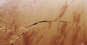 An aerial view of Mars' surface featuring a large, dark crack running horizontally across the center. The terrain is predominantly reddish-brown, with varying textures and streaks spread throughout. A scale indicator at the bottom-right shows 20 kilometers.
