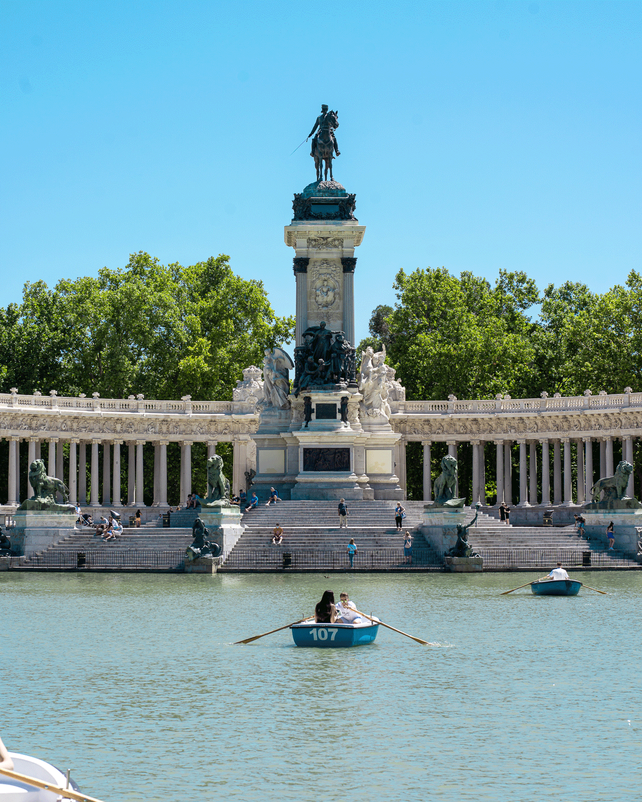 Image shows a large stone monument featuring a tall pillar topped by a statue of a man on horseback, located in a serene park. In the foreground, people are rowing boats on a reflective lake. The background includes a colonnade and lush green trees under a clear blue sky.