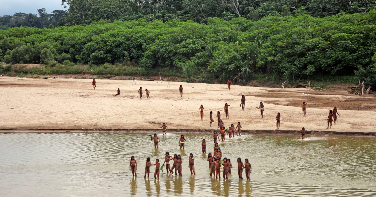 A group of people, likely indigenous, are gathered along a sandy riverbank and wading in shallow water. The background is lush with dense green foliage. Some individuals are in the water, while others are scattered along the shore.