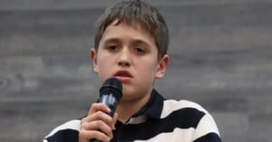 A young person with short brown hair is holding a microphone and speaking. They are wearing a black and white striped shirt and have a serious expression. The background is a gradient of gray tones.