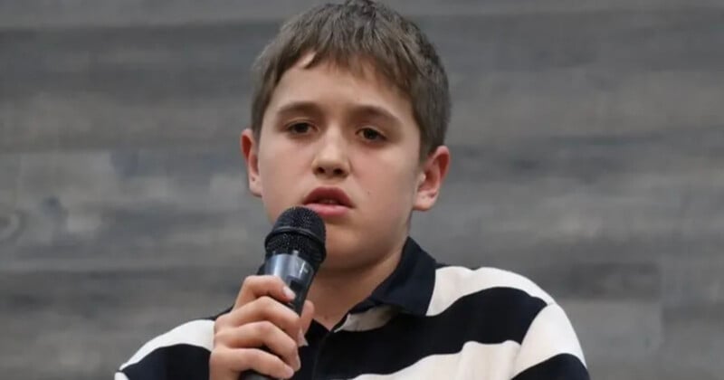 A young person with short brown hair is holding a microphone and speaking. They are wearing a black and white striped shirt and have a serious expression. The background is a gradient of gray tones.