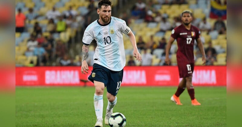 A soccer player wearing a white and blue striped jersey with the number 10 dribbles the ball on a green field. A player in a maroon uniform is visible in the background. The scene is set in a stadium filled with spectators, with the stands and advertisements blurred.