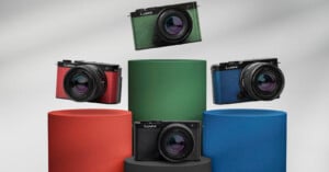 Four Lumix cameras with different body colors (red, green, blue, and black) are displayed on colorful cylindrical pedestals (orange, green, blue, and black) against a neutral background. Each camera has a large lens, and the arrangement showcases the variety available.