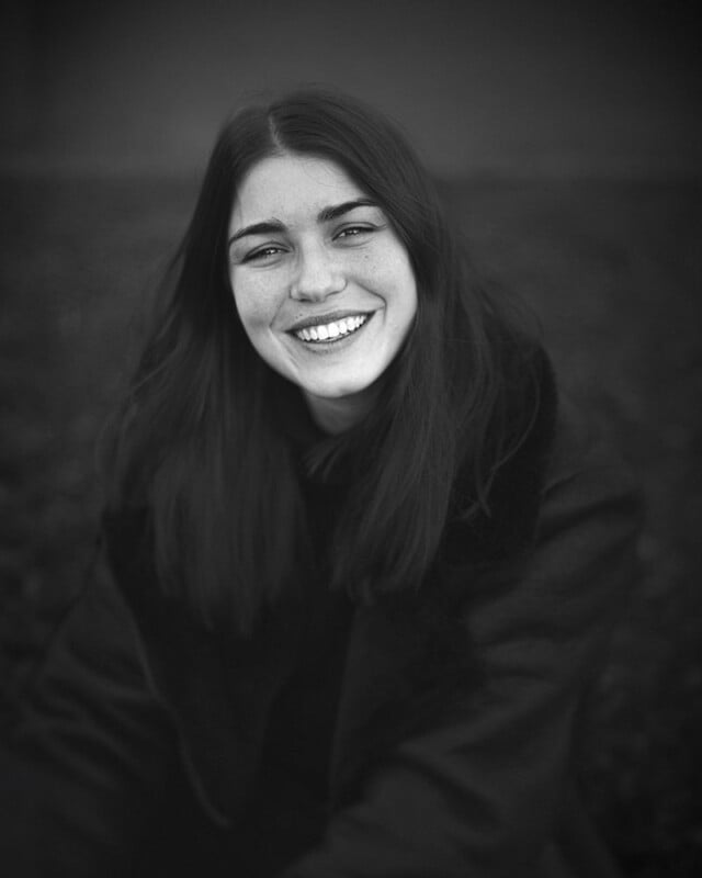 A black and white photo of a young woman with long dark hair, smiling broadly. She is wearing a dark coat with a fur collar and looking directly at the camera. The background is out of focus, emphasizing her cheerful expression.