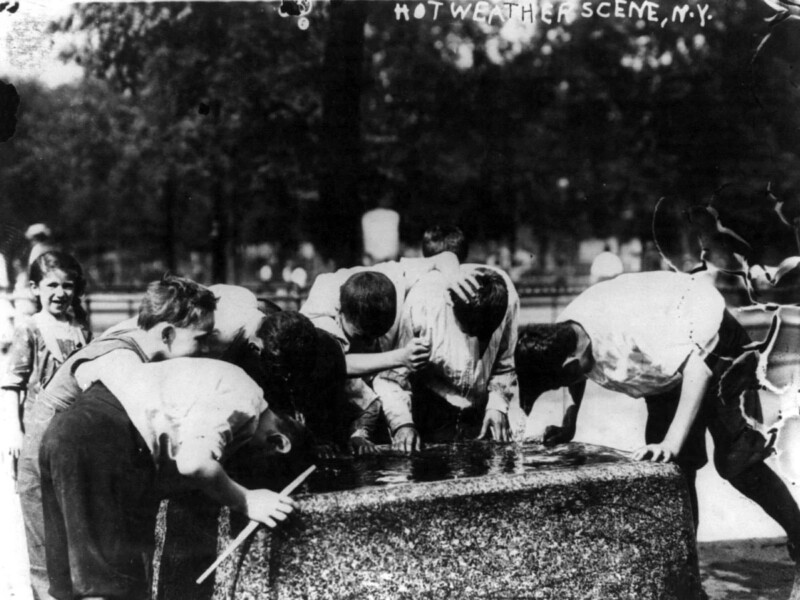 A black and white photo titled "Hot Weather Scene, N.Y." depicts a group of boys leaning over a water fountain, drinking or washing their heads. They appear to be cooling off on a hot day in New York. Trees and blurred figures are visible in the background.