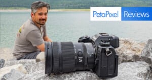 A man with short hair and a beard sits on rocks by a lake, looking at the camera, with greenery in the background. In the foreground is a Nikon camera with a large lens. A banner reads "PetaPixel Reviews.