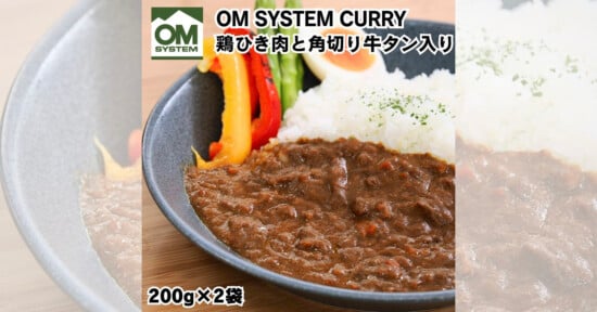 A plate of dark brown curry with beef chunks and chicken is served with white rice and colorful bell pepper slices. The text states "OM SYSTEM CURRY" with package information "200g x 2". The OM SYSTEM logo is in the top left corner, with text in Japanese.