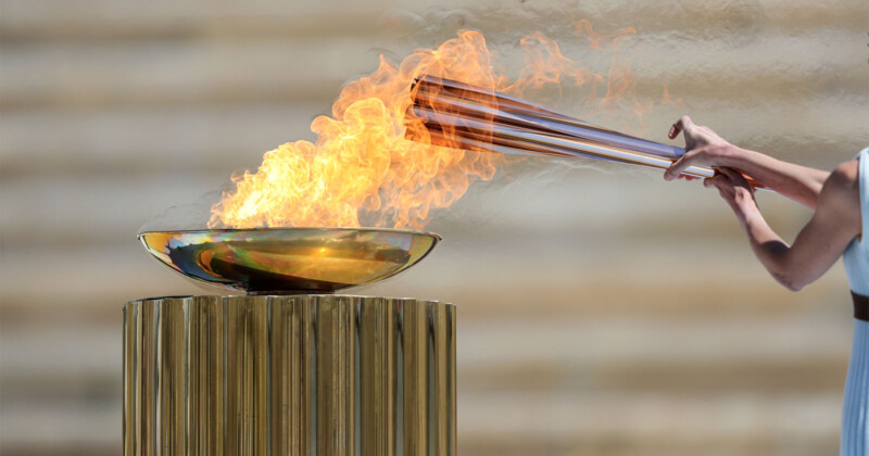 A person lighting a large ceremonial flame with a torch in a gold-colored bowl on a pedestal. The torchbearer's face is not visible, and they are dressed in light-colored attire. The background is blurred and neutral, putting focus on the flaming ceremony.