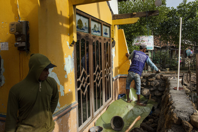 Two construction workers repair a stone structure next to a yellow-painted house. One worker in a green hoodie stands near the wall, while the other works with stones in a blue shirt. The house has a large window with decorative bars and plants around it.