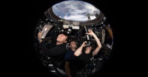 Three astronauts inside the International Space Station's cupola, capturing images of Earth using cameras. The window offers a striking view of the planet's surface. They appear focused, with one pointing and the others looking through their cameras.