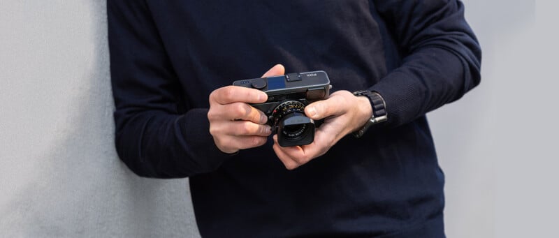 A person wearing a navy blue sweater and a black watch holds a compact camera with both hands, positioned near their midsection, against a gray background.