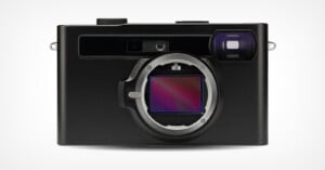 A modern black digital camera viewed from the front. The lens is removed, revealing the camera's sensor inside the mount. The camera has a rectangular design with rounded edges and a viewfinder on the top right. The background is plain white.
