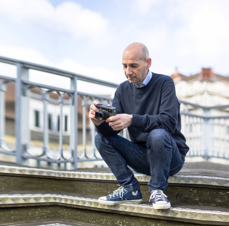 A bald man in a navy blue sweater and jeans crouches on outdoor steps, holding a camera. He appears to be examining or adjusting the camera settings. The background features a metal railing and blurred buildings. The sky is clear with a few clouds.