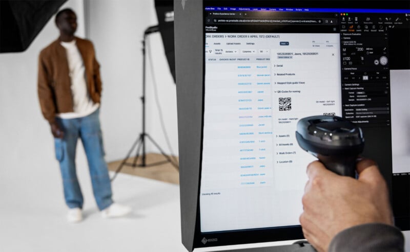 A person in casual attire is blurred in the background while a hand in the foreground holds a barcode scanner in front of a computer screen displaying shipping information. The setting appears to be a studio with professional lighting equipment.