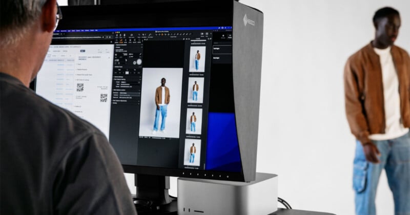 A person wearing a brown jacket and blue jeans is modeling in front of a white background. Another person is operating a computer, reviewing photos of the model displayed on a monitor.