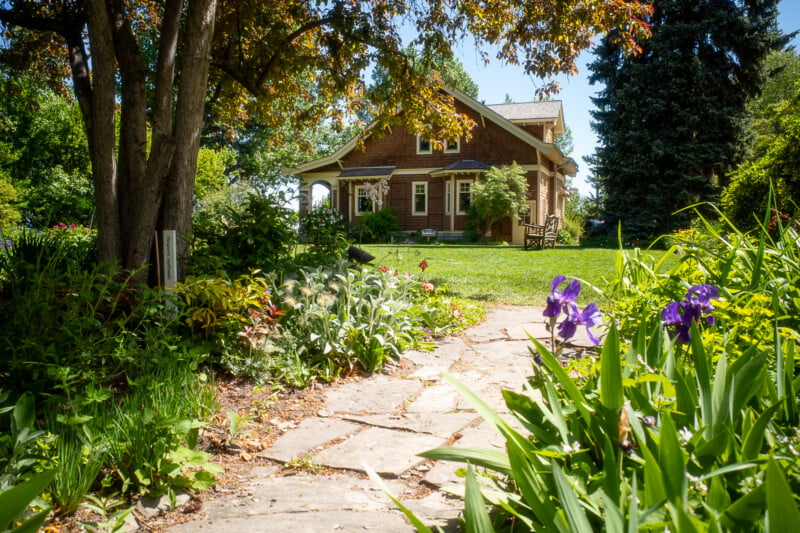 A stone path leads to a charming brown house surrounded by lush greenery and vibrant flowers, including purple irises, under a clear blue sky on a sunny day. Tall trees provide shade, and a wooden chair sits on the well-maintained lawn.