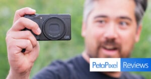 A person holds up a black Ricoh GR camera in their left hand, displaying it prominently. The background is blurred green foliage. In the bottom right corner, there's a banner with the text "PetaPixel Reviews." The person is smiling and partially out of focus.