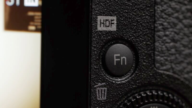 Close-up shot of a camera's back panel showing three buttons: a grey, square "HDF" button at the top, a round black "Fn" (Function) button in the center, and a grey, square button with a trash bin icon at the bottom, all against a textured black surface.