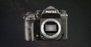 Front view of a Pentax K-1 camera body against a starry background. The camera's lens is not attached, revealing the sensor inside. The camera has a textured grip with control buttons visible on the body.