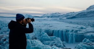 A person wearing a dark jacket and a blue beanie takes a photo with a camera while standing in an icy, mountainous landscape at dusk. The scene includes towering ice formations and a vast, frozen expanse.
