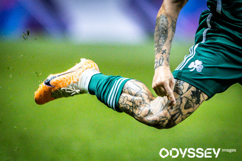 A close-up of a soccer player's tattooed leg wearing green shorts and an orange cleat in mid-air during a game. The background is blurred, focusing attention on the player's dynamic movement and detailed tattoos. The image is by ODYSSEY Images.