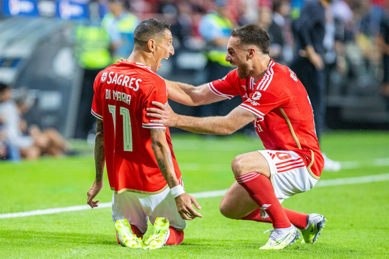 Two soccer players wearing red jerseys celebrate on the field. The player on the left, with "DI MARIA 11" on his back, is kneeling and smiling, while the player on the right is crouched, reaching out with both hands to touch his shoulders. The background shows spectators and other players.