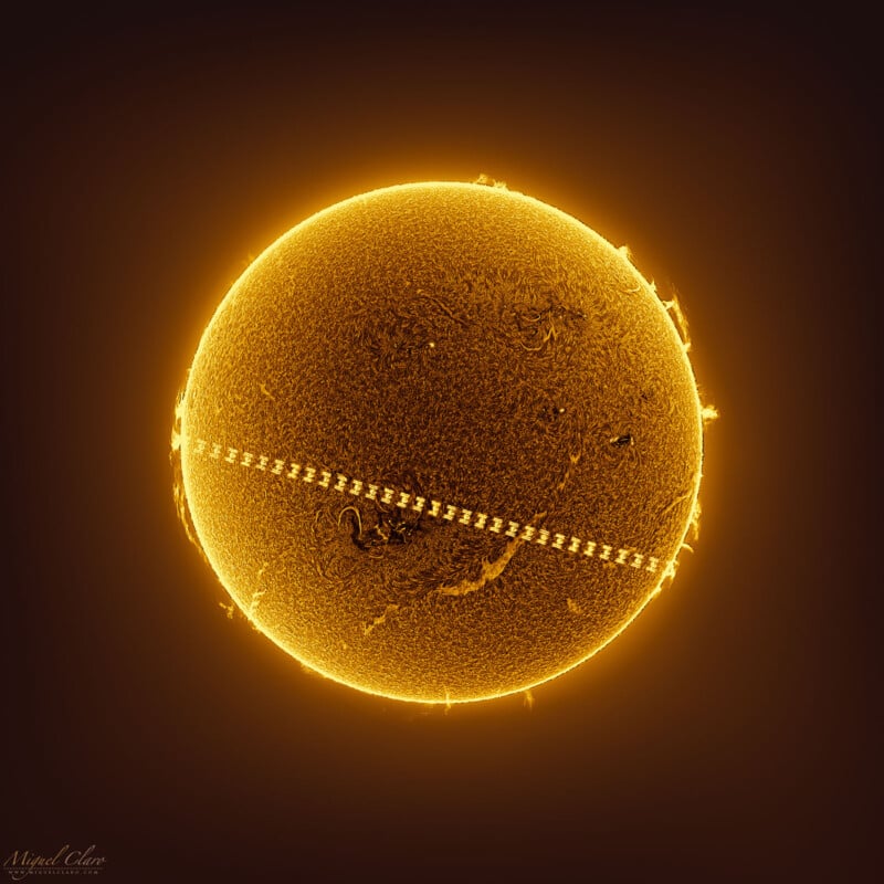 Una imagen del sol con una apariencia brillante de color amarillo dorado sobre un fondo oscuro.  La superficie está detallada con textura visible y destellos solares.  Una línea diagonal de puntos cruza el Sol y representa el tránsito de la Estación Espacial Internacional (ISS).