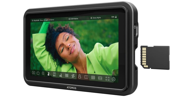 A black Atomos video monitor displays the image of a person smiling with closed eyes, wearing a green blazer and lying on green grass. An SD card is partially inserted into the monitor's side slot. The monitor screen shows various control icons at the bottom.