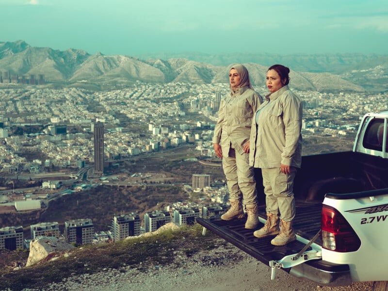 Two women in beige outfits stand on the tailgate of a white pickup truck, overlooking a sprawling cityscape nestled among mountains. The sky is clear, and the city, with modern buildings and open spaces, extends towards the horizon. The women look resolute.