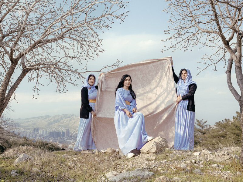 Three women stand in a serene outdoor setting with bare trees and a distant city skyline. The central woman, dressed in a light blue gown, sits on a rock, while the other two hold up a large piece of light fabric behind her, creating a modest backdrop.