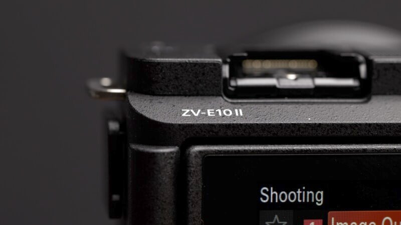 Close-up of a black camera with the inscription "ZV-E10II" above. The camera screen shows part of the user interface with the word "Shoot" visible on a dark background. Details of the buttons and the design of the camera are partially visible.