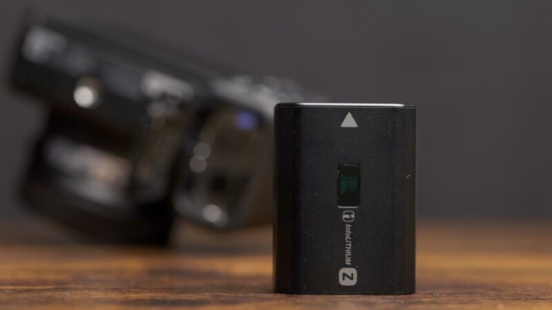 Close-up of a black lithium-ion battery standing upright on a wooden surface. The battery features a small green window and a white arrow pointing upwards. In the background, a blurry camera can be seen against a dark, blurry background.