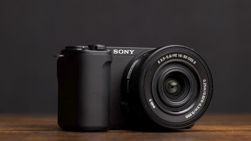 A close-up of a black Sony mirrorless camera with a 16-50mm lens attached. The camera is set against a dark background and resting on a wooden surface. The lens details are clearly visible, showing the zoom range and aperture information.