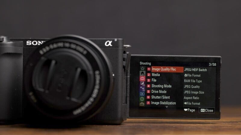 A Sony α camera with the LCD screen unfolded to the right. The screen displays menu settings, including options for image quality/recording, media, file, recording mode, drive mode, shutter/silent, and image stabilization.