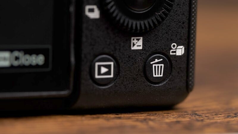 Close-up of the rear control buttons on a camera. Visible buttons include a play button with a video playback icon, a delete button with a trash can icon, and other function buttons. The surface looks like the corner of a camera body resting on a wooden surface.
