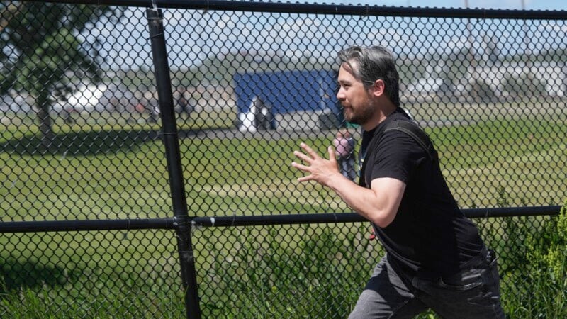 A man with grey hair and a beard wearing a black shirt and grey pants is seen mid-jog next to a chain link fence on a sunny day. A grassy field and a tree can be seen in the background.