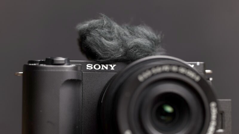 Close-up of a Sony camera, focusing on the lens and brand logo. There is a blurred windscreen attached to the top of the camera, partially covering the built-in microphone. The background is blurred, highlighting the details of the camera.