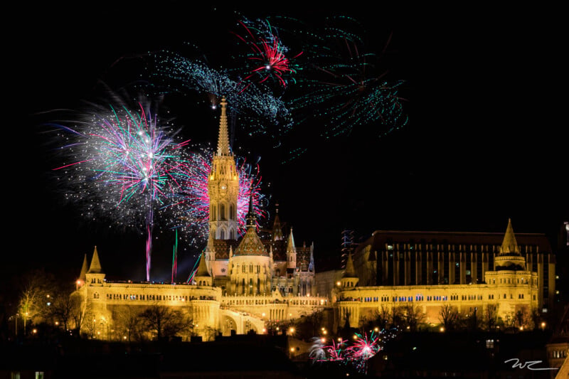 A spectacular display of colorful fireworks illuminates the night sky above the beautiful Gothic-style building with its tall ironwork and beautiful architecture.  The surrounding buildings are also illuminated, which adds to the atmosphere.