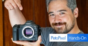 A person with gray hair and a beard smiles and points to a Canon EOS R5 camera with the lens removed, showcasing the camera sensor. The image has a "PetaPixel Hands-On" logo in the bottom left corner.