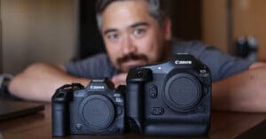 A person with blurred facial features sits in the background, while two Canon EOS cameras, one smaller model (EOS R5) and one larger model (EOS-1D), are prominently displayed in the foreground on a wooden surface.