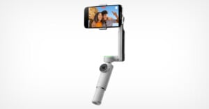Photo of a white handheld smartphone stabilizer with an attached smartphone. The phone displays an image of two individuals smiling and waving. The device has a circular camera at the base, designed to aid in capturing steady video and photos.