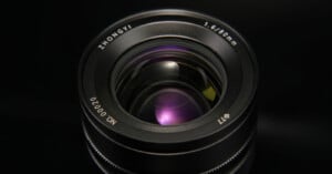 Close-up view of a camera lens with visible details such as the brand name "ZHONGYI," 1.6/60mm, lens serial number "NO:00020," and filter size "Φ77." The glass reflects a purple hue, set against a dark background.