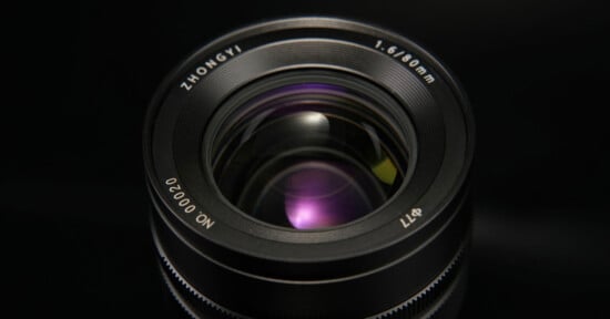 Close-up view of a camera lens with visible details such as the brand name "ZHONGYI," 1.6/60mm, lens serial number "NO:00020," and filter size "Φ77." The glass reflects a purple hue, set against a dark background.